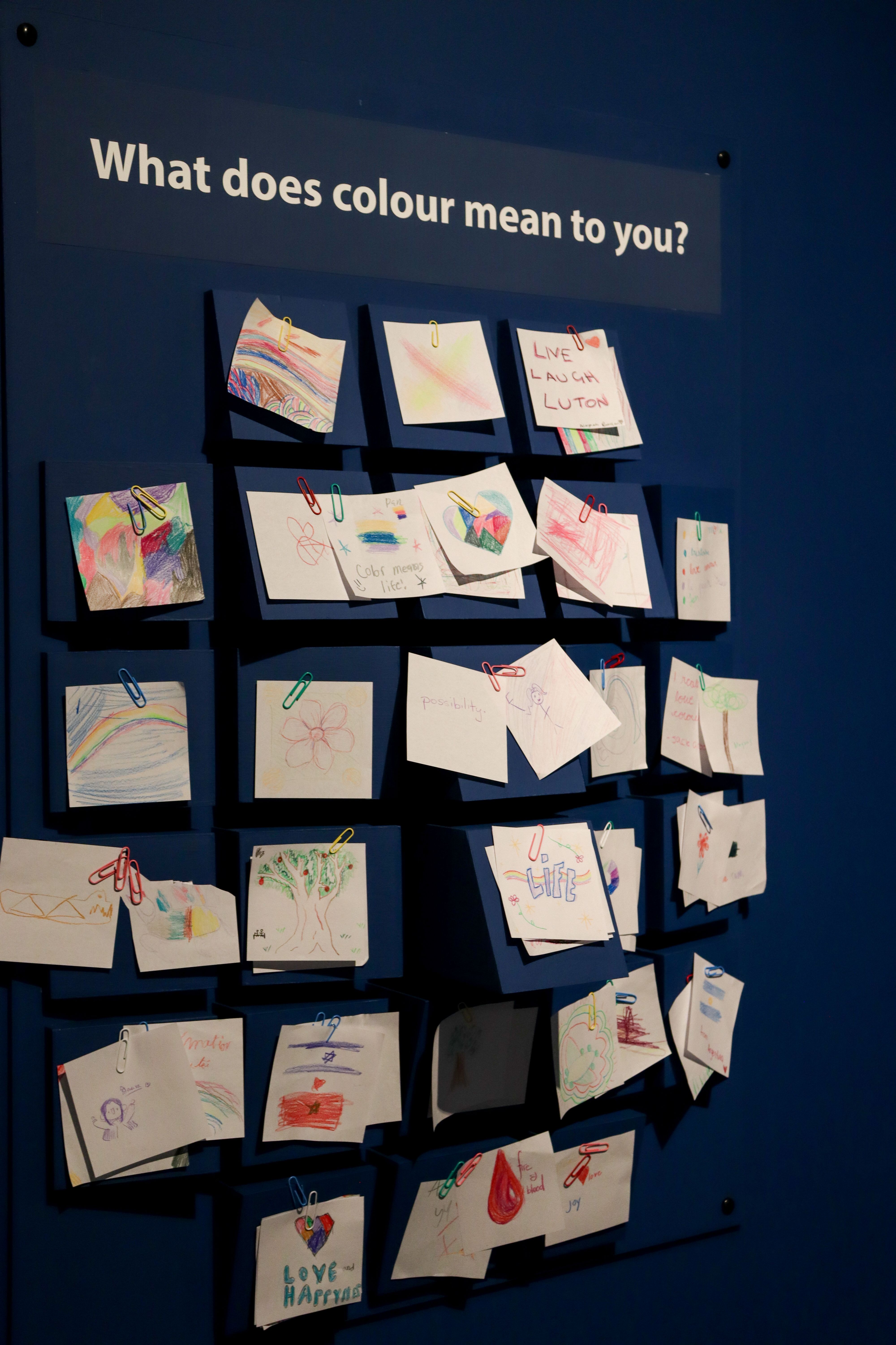 An installation against a blue wall, with the words 'What does colour mean to you?' at the top. It resembles a face, with many square boxes that people have stuck post it notes onto with magnetic clips. The notes have drawings, notes, and comments about colour all over them.