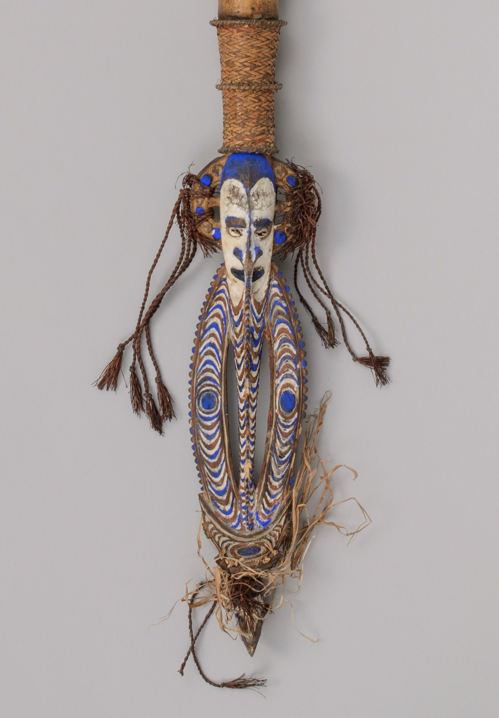 Bamboo tube with wooden ornament in shape of a janus-face at top of barred ovoid, with a bird on a ' nose' at the ovoid's tip. Decorated with blue. white and red paint and many tassels. Photographed against a grey background.