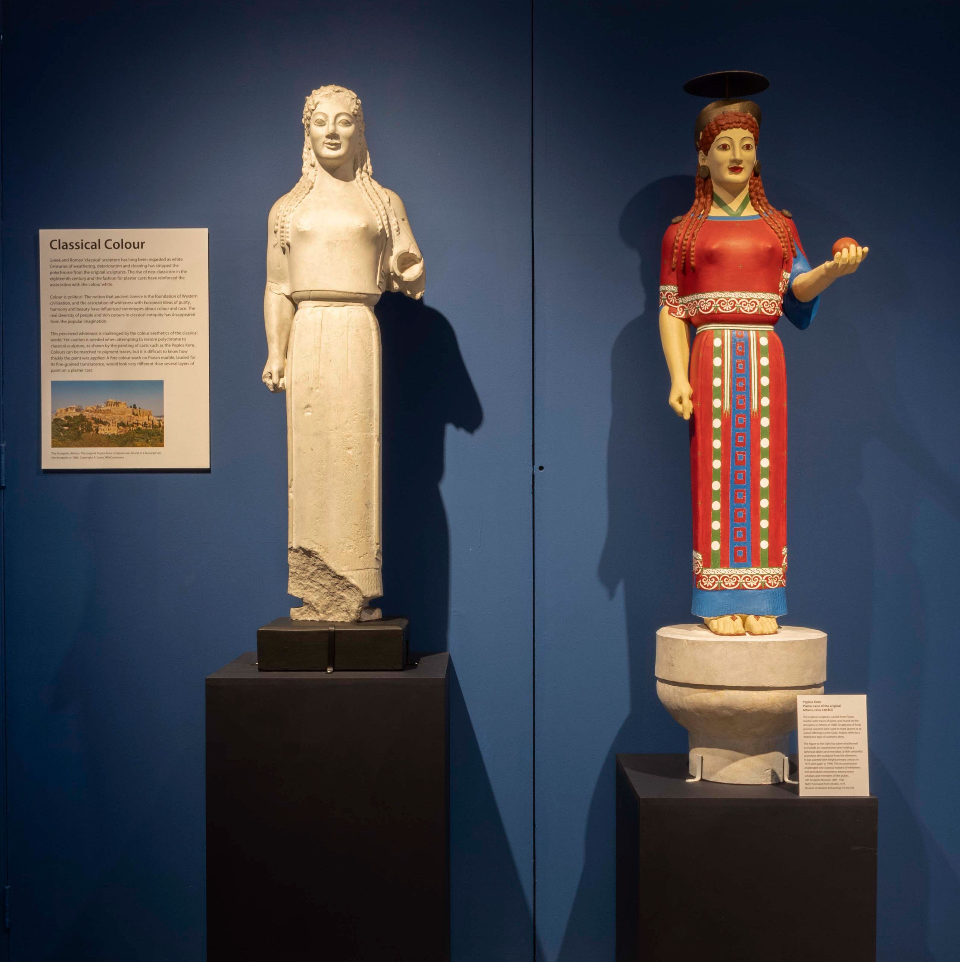 Two statues standing in a gallery, against a blue wall. They are both of a woman in the same position, one arm down and one held up, holding an apple. She wears a long tunic dress and has long, curled hair. The left statue is made of white stone, whilst on the right, slightly smaller in size, she is painted in vibrant colours; a red dress with blue, green and white detailing, red hair, and gold headdress and earrings. 