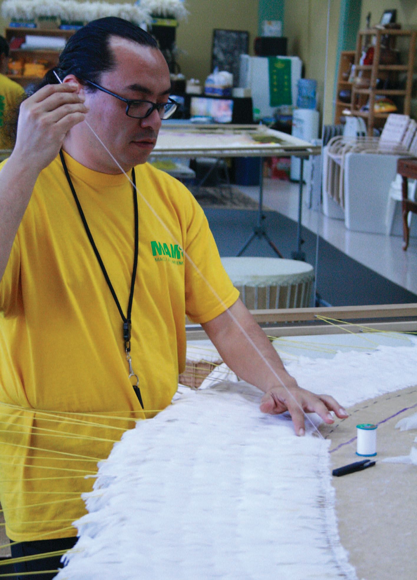 A man with black hair, held back in a ponytail, and wearing glasses and a white t-shirt and black lanyard, stitching feathers onto a long burlap background. It looks like a long, white, neatly fluffy piece.