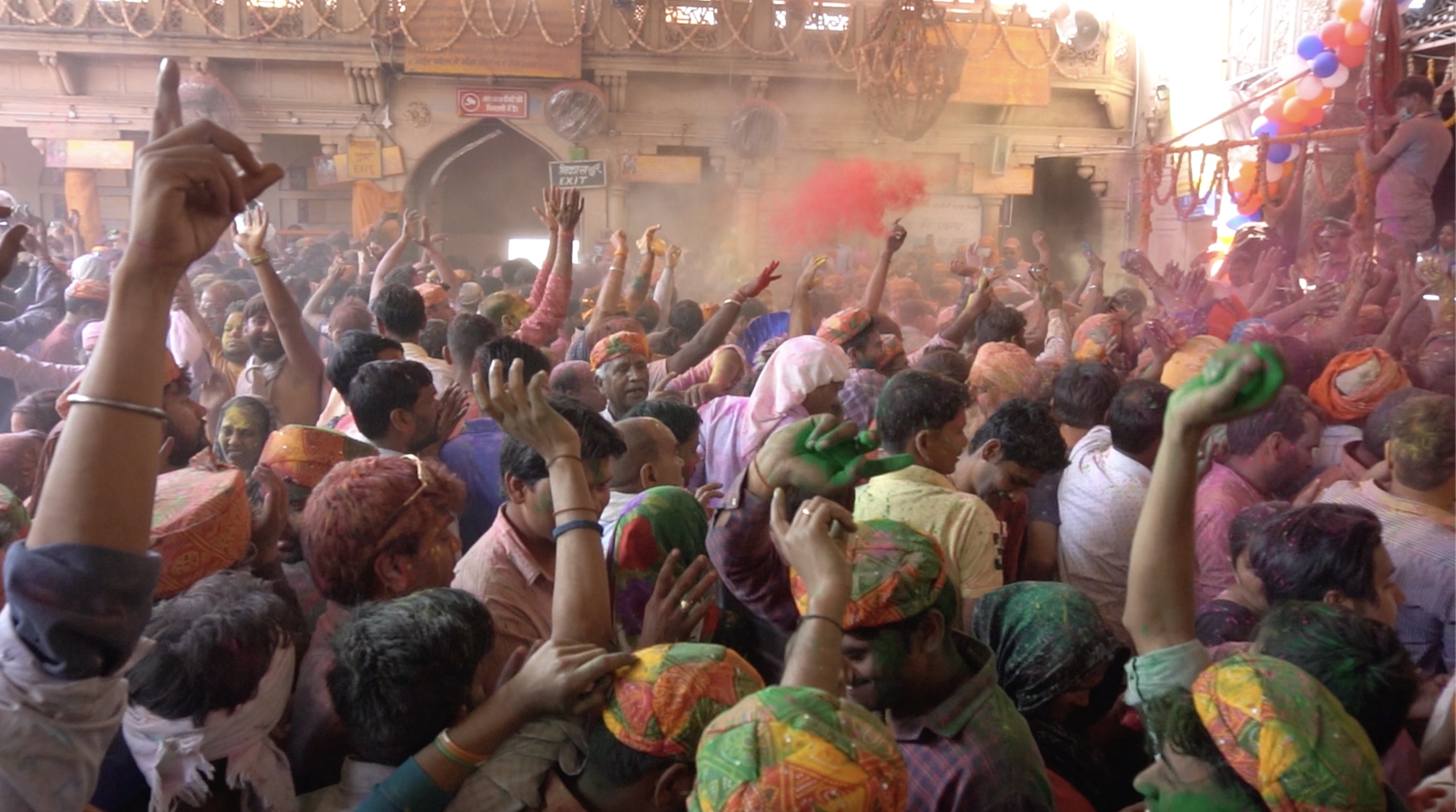 Holi festival in Barsana, India. The festival is very busy, with people dancing with their hands in the air, and colour pigments being thrown around. Some people are already covered in pigmented powder. The air is dusty, and a stage is visible on the right.