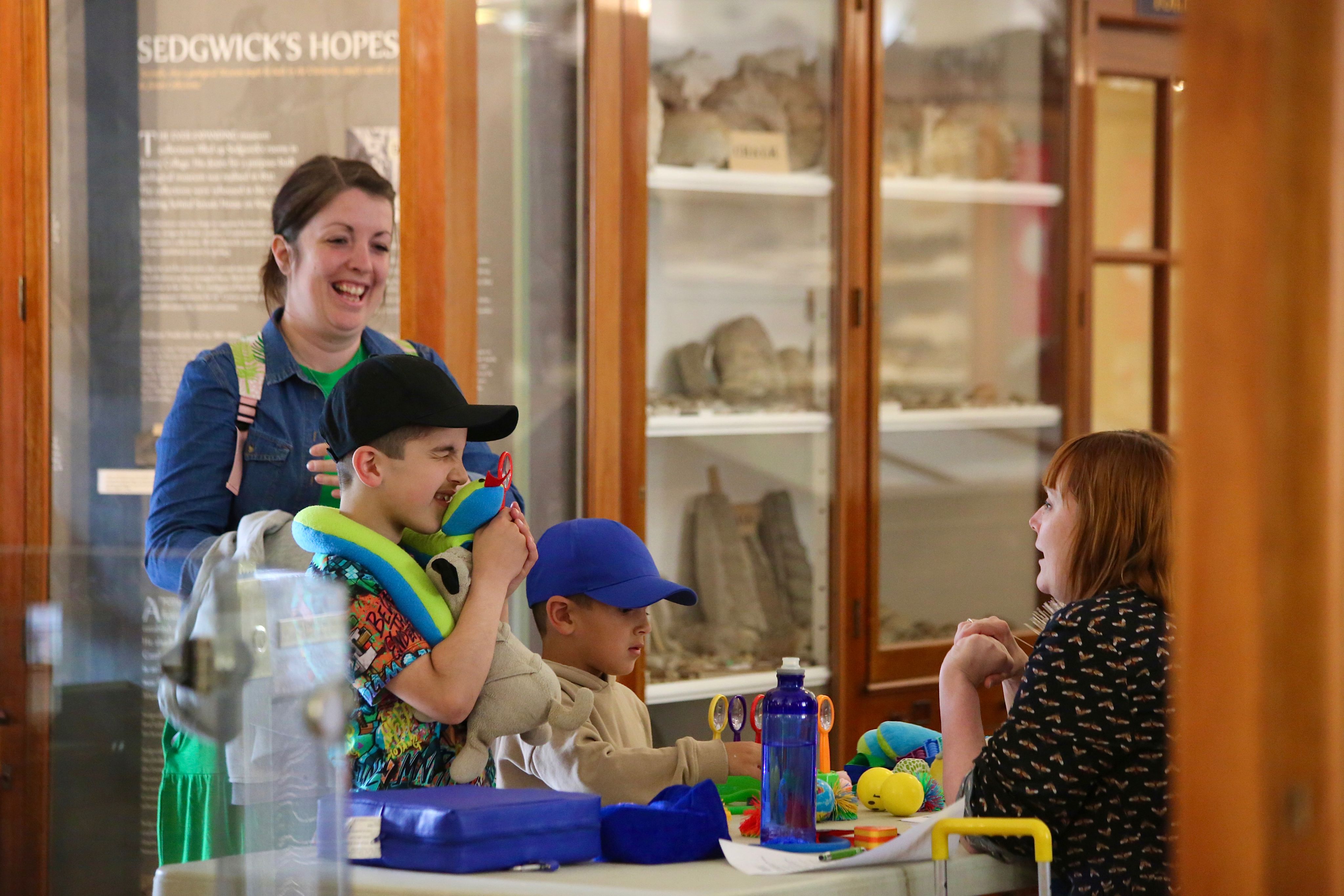 A woman stands with two boys in front of a desk at the Sedgwick Museum that is filled with sensory toys. One boy grins and holds a toy up to his face. You can see wooden display cases in the background.