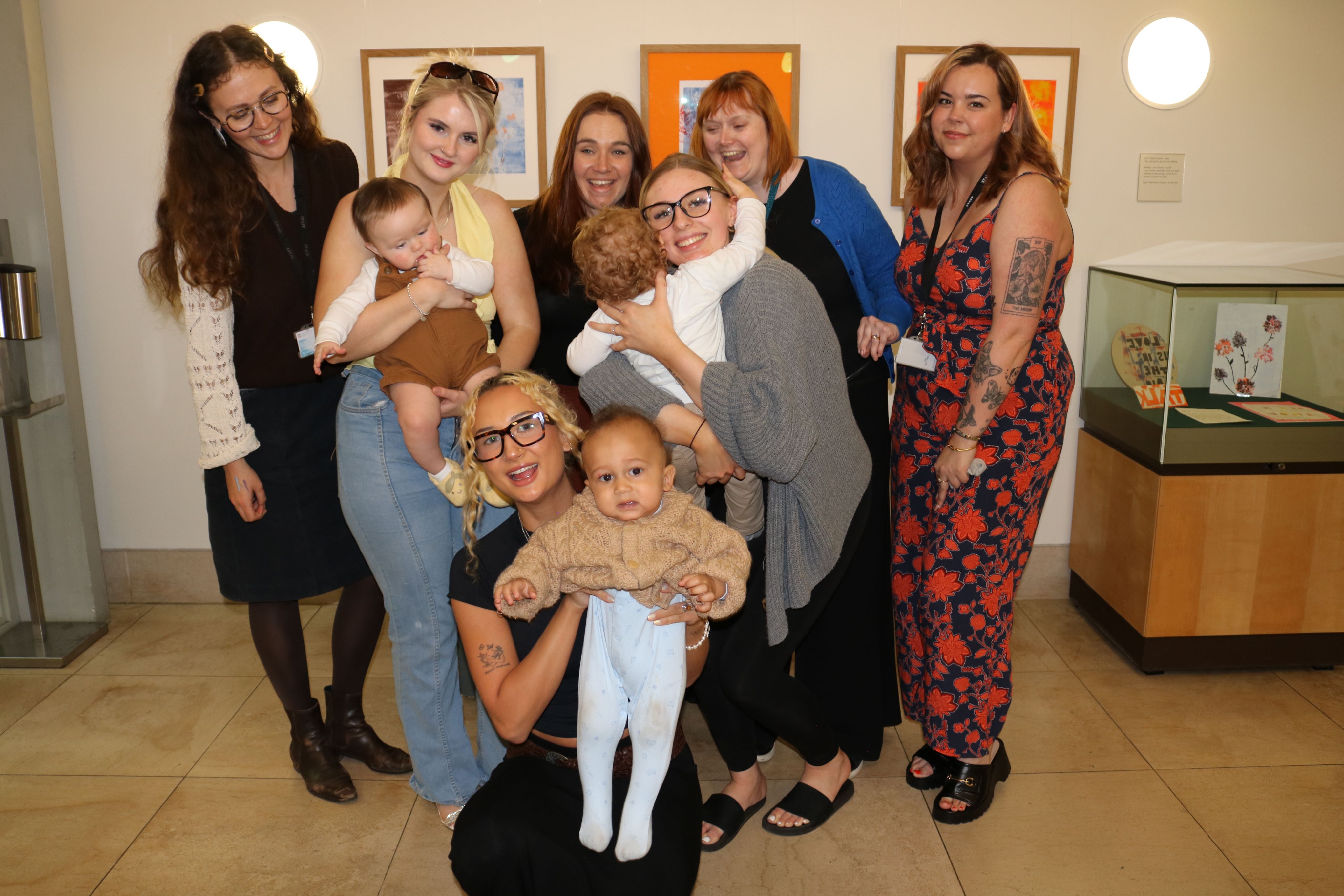A group of seven adults stand smiling in a group in front of their exhibition at the Fitzwilliam Museum, three holding their children in their arms.