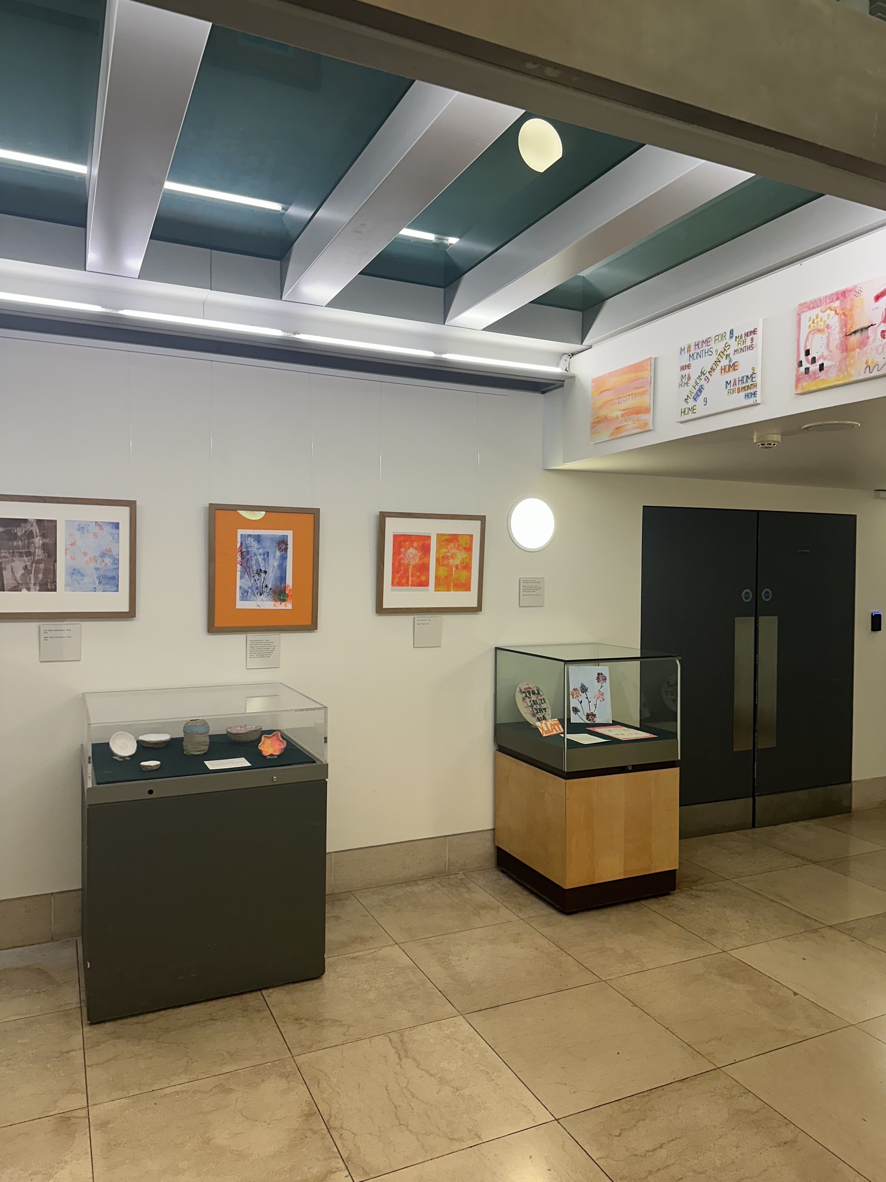 The Young Parents exhibition at the Fitzwilliam Museum. two glass display cases are filled with artworks and three framed artworks are mounted on the walls with labels. Three other canvases are up on a higher adjacent wall.