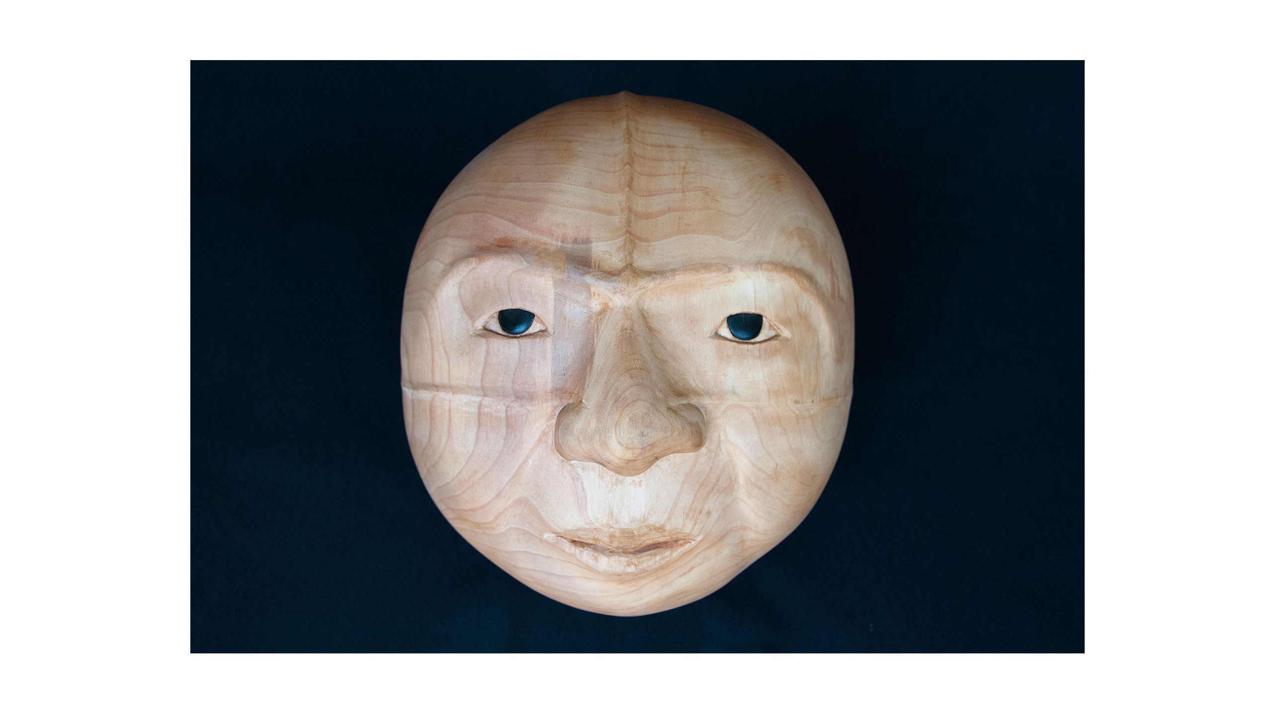 Wooden carving of a round face, with dark eyes made of baleen and a prominent brow reminiscent of a whale tail.