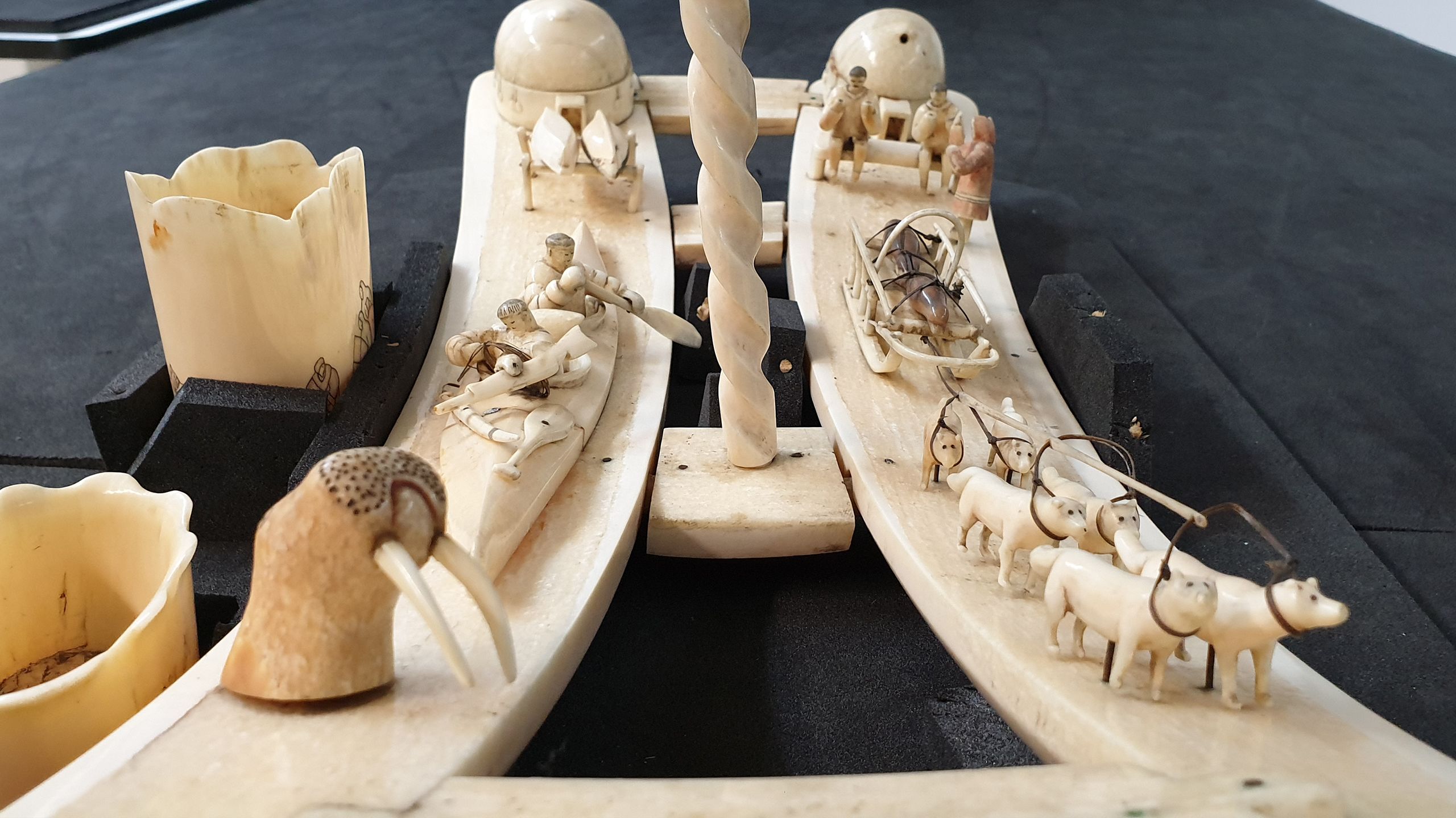 Walrus ivory carving featuring walrus, huskies pulling a sledge which in turn has seals loaded onto it, a kayak and some figures in the background.