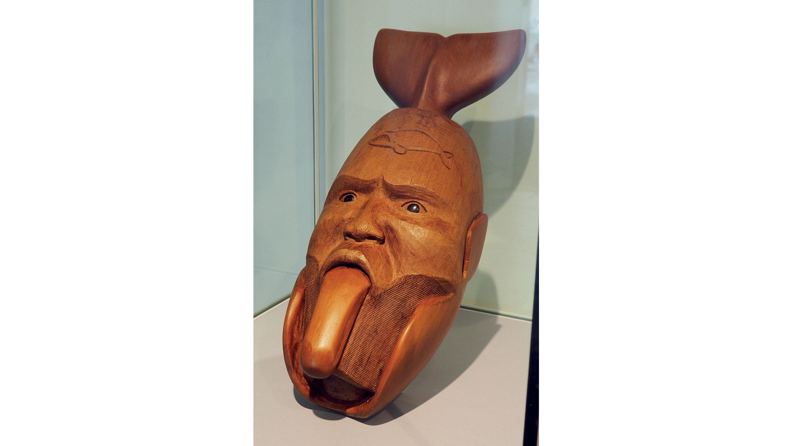 Wooden mask carving depicting a head in the process of transforming from a human face to a whale, with a whale tail protruding from the top of the head.